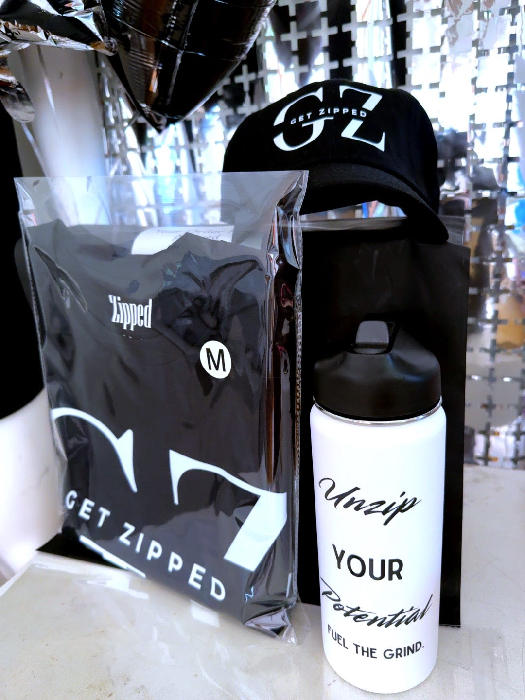 Zipped Lifestyle Starter Bundle: black ‘Get Zipped’ tee in poly bag (size M), black GZ dad hat, and white bottle reading ‘Unzip Your Potential — Fuel the Grind