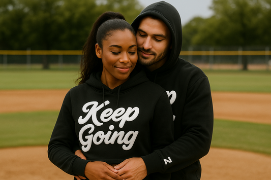 Young couple in black “Keep Going” hoodies with Z logo on sleeve, embracing on a baseball field.